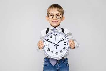 A charming boy in a white shirt, suspenders, a tie and light jeans stands on a gray background. The...