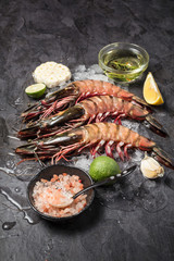 Three raw jumbo shrimps on ice with basic ingredients to cook on dark background. Vertical composition