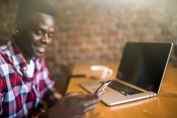 Handsome young Afro American smiling happily while reading sms on smart phone, messaging using free wi-fi, having coffee at cafe