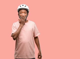 Black man wearing bike helmet covers mouth in shock, looks shy, expressing silence and mistake concepts, scared
