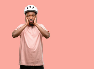 Black man wearing bike helmet covering ears ignoring annoying loud noise, plugs ears to avoid hearing sound. Noisy music is a problem.