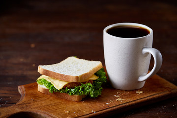 Breakfast table with sandwich and black coffee on wooden cutting board over rustic background, close-up, selective focus