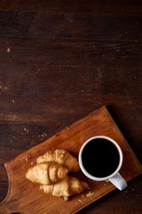 Breakfast with croissants and black coffee on wooden cutting board over rustic background, close-up, selective focus