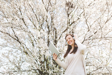 Naklejka premium Young smiling beautiful woman in light casual clothes with headphones listening music holding tablet pc computer in city garden or park on blooming tree background. Spring flowers. Lifestyle concept.