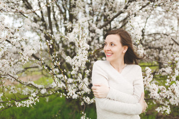 Young smiling happy charming woman in light casual clothes holding hands folded standing in city garden or park on blooming tree background. Spring nature, flowers. Lifestyle, leisure concept.