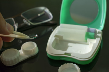 Fototapeta na wymiar contact lens in tweezers on the background of glasses, a green lens container and a solution with a mirror lying on a table