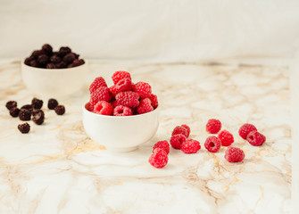 Raw raspberries and wild black, blackberries in white cups. Light marble background. Summer fruit, berries. Detox diets and healthy food concept.