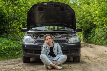A young girl sits near a broken car on the road with an open hood and calls the rescue service to...