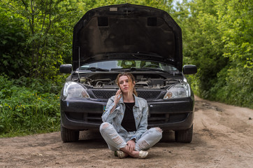 A young girl sits near a broken car on the road with an open hood and calls the rescue service to get her car repaired