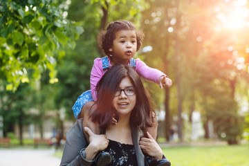 Mixed race mother carrying excited hispanic toddler daughter three years old in blue denim sarafan and pink shirt holding on her shoulders while walking together in summer park.