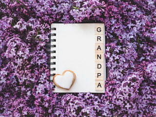 Heart-shaped cookie, covered with bright glaze, word GRANDPA, notepad with a blank page for Your inscription on the background flowers. Top view, close-up. Congratulations for relatives, loved ones