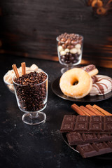 Glass with coffee beans next to chocolate tablets, donuts, brown sugar and other glass with peanuts in chocolate on dark wooden background