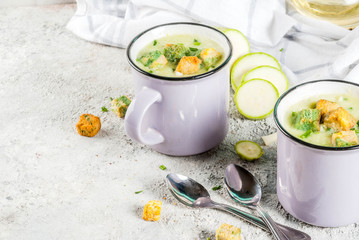 Two portions of homemade zucchini creamy soup with bread crumbs in mugs on a light concrete background.