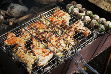 Juicy chicken wings on barbecue grill and mushrooms on metal skewers are being prepared over charcoal. Bbq meat and grilled vegetables, also called shish kebab or shashlik, close up