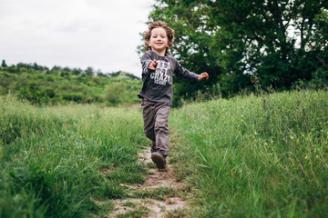 Naklejka na ściany i meble Little boy running down a meadow in a beautiful landscape in summer, very light and happy scene. Curly toddler