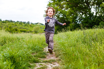 Little boy running down a meadow in a beautiful landscape in summer, very light and happy scene. Curly toddler