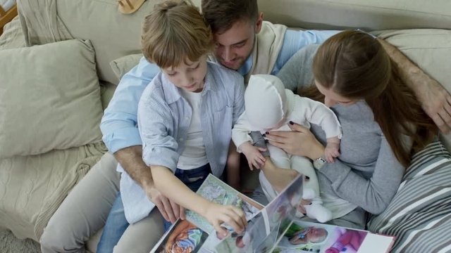 Top view of happy young parents with elementary age son and baby boy sitting on sofa and looking at photographs in album