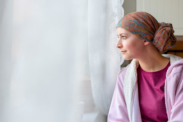 Young adult female cancer patient wearing headscarf and bathrobe sitting in the kitchen looking out...
