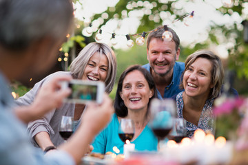 Some friends in their forties gathered around a table in the garden for a good time. A man takes a picture of a group of friends