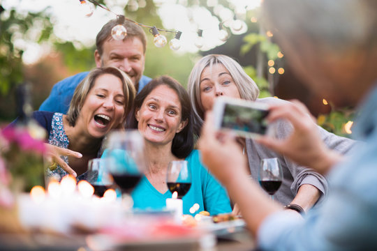 Some friends in their 40s gathered around a table in the garden to share a meal. A man takes a picture of a group of friends