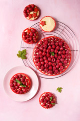 Variety of red raspberry shortbread tarts and tartlets with lemon custard and glazed fresh raspberries served on cooling rack over pink pastel background. Top view, space.