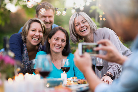 Some friends in their forties gathered around a table in the garden for a good time. A man takes a picture of a group of friends