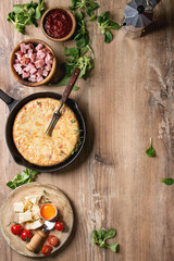 Spanish potato omelette tortilla with bacon served in cast-iron pan with sauce and ingredients above over wooden background. Top view, space.
