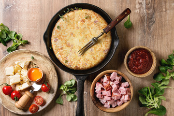 Spanish potato omelette tortilla with bacon served in cast-iron pan with sauce and ingredients above over wooden background. Top view, space.