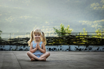 Fototapeta na wymiar a little girl with long blonde hair and a charming smile sitting in the Lotus position and meditates on a background of mountains at dawn