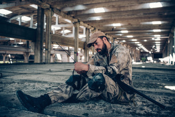 Soldier is sitting on the ground and looking at his portable radio. He is holding it with both hands. Guy has curved his left hand and out it under right one. He has rifle lying on his left leg.