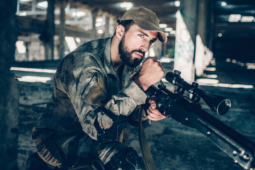 Calm and peaceful soldier is looking straight forward. He is waiting. Young man is sitting one one knee. Guy wears military uniform.