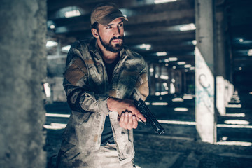 Fototapeta premium Calm fighter wears military uniform. He is standing besides column and paying good attention. He is watching. Guy has a black pistol in his hands. Also man has a finger on trigger.