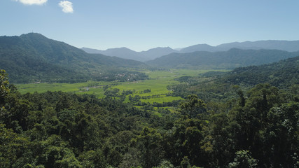 Fototapeta premium Mountain valley with village, farmland, rice fields. Aerial view of Mountains with green tropical rainforest, trees, jungle with blue sky. Philippines, Luzon.