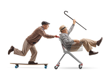 Senior riding a longboard and pushing a shopping cart with another senior riding inside it