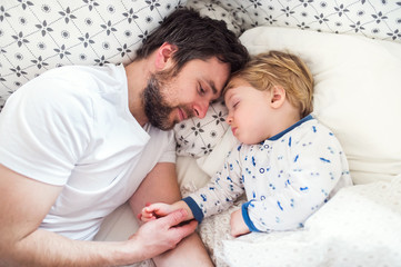 Father holding hand of a sleeping toddler boy in bed at home.