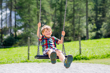 Funny kid boy having fun with chain swing on outdoor playground while being wet splashed with water
