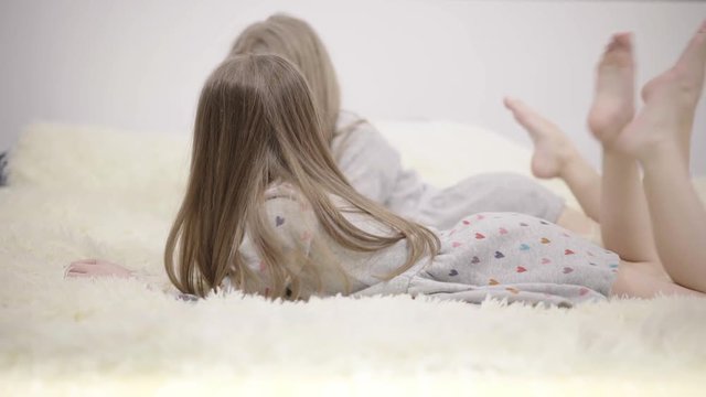 Two little girls with long fair hair wearing gray dresses are lying on a fluffy bed cover and talking. Concept of a happy family and carefree childhood. Locked down real time medium shot