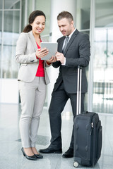 Full length portrait of successful business couple, man and woman, standing in hall of modern airport with suitcase and using digital tablet