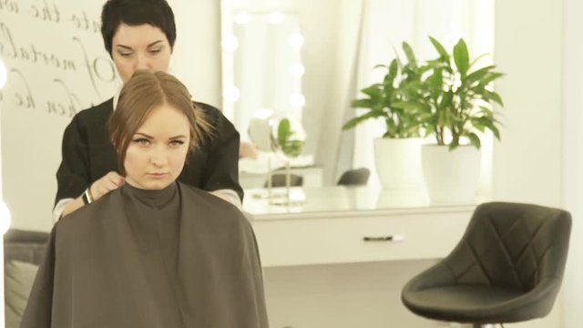 Hairdresser wearing cloack robe on young woman before haircut in hair studio. Preparing for haircut in hairdressing salon front mirror.