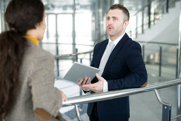 Waist up portrait of modern businessman holding laptop talking to female colleague standing by railing in office building, copy space