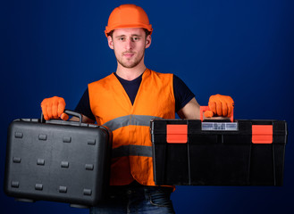 Man in helmet, hard hat holds toolbox and suitcase with tools, blue background. Worker, repairer, repairman, builder on thoughtful face choosing equipment for work. Toolbox and equipment concept.