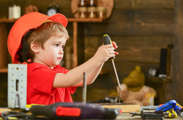 Side view little kid binding screw into wooden block. Concentrated boy in orange helmet sitting at...