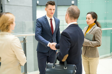 Portrait of handsome young businessman shaking hands with partner after meeting on beneficial deal standing with group of colleagues in hall of modern office building