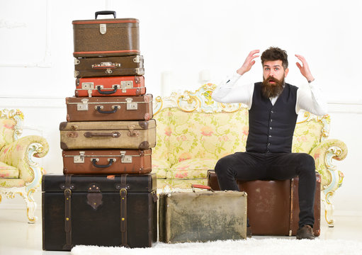 Macho elegant on surprised face sits shocked near pile of vintage suitcase. Luggage and relocation concept. Man, butler with beard and mustache delivers luggage, luxury white interior background.