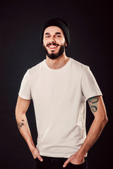 Waist up portrait of serious stylish attractive man with thick beard looks confidently, dressed in casual white t shirt, isolated over black background. Young hipster in cap with tattoo. Mock up
