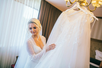 Beautiful blonde bride in robe posing near white wedding dress.  Morning of the bride. Woman in...