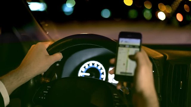 Unrecognizable man driving his car in a night city and web surfing with his phone. Concept of dangerous driving behavior and recklessness. Handheld real time medium shot