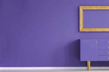 Real photo of a creative living room interior with golden frame above violet cupboard standing...