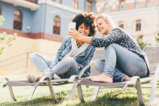 Two young beautiful laughing girls of different races are sitting outdoors on the recliners on the lawn and taking the selfie using the smartphone; interracial female couple photographing on cellphone