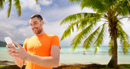 fitness, sport and technology concept - smiling man with smartphone and earphones listening to music over tropical beach background in french polynesia